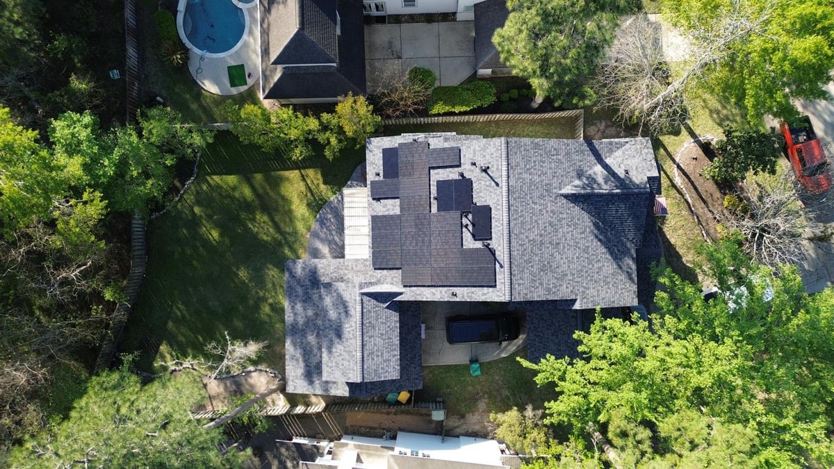 Aerial view of a modern house with gray shingle roof surrounded by green trees and neighboring properties