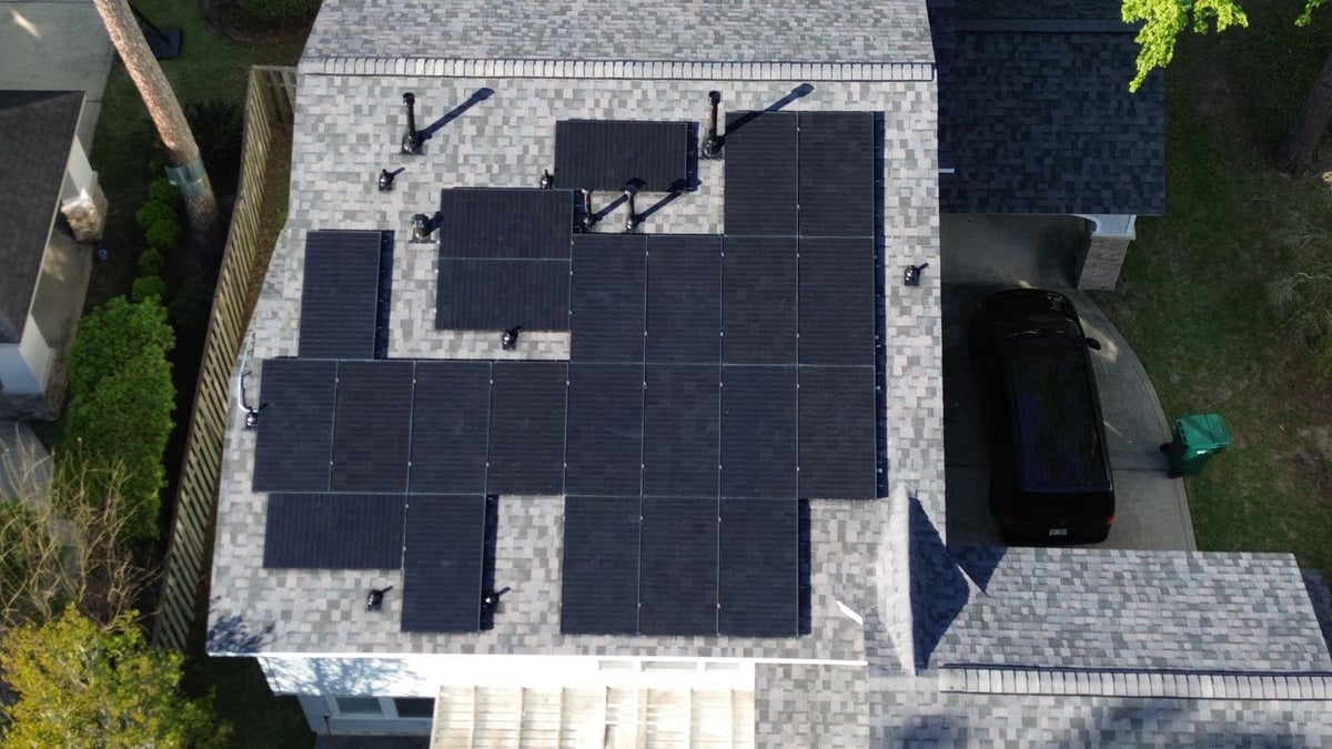 Aerial view of residential roof with installed solar panels arranged in a pattern, surrounded by landscaping and adjacent buildings.