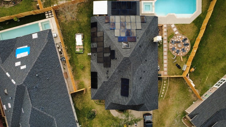 Aerial view of a residential house with dark gray roof surrounded by green lawn and neighboring properties with pool visible