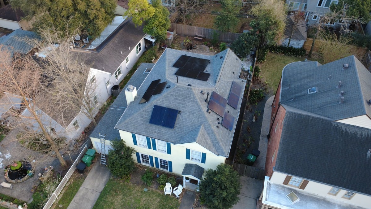 Aerial view of a blue and white residential house with dark roof and solar panels, surrounded by trees and neighboring homes