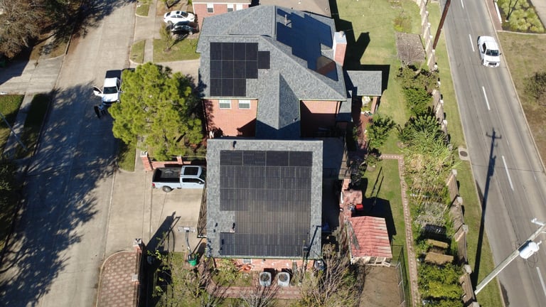 Aerial view of residential house with solar panels on roof, driveway, and surrounding trees and landscaping