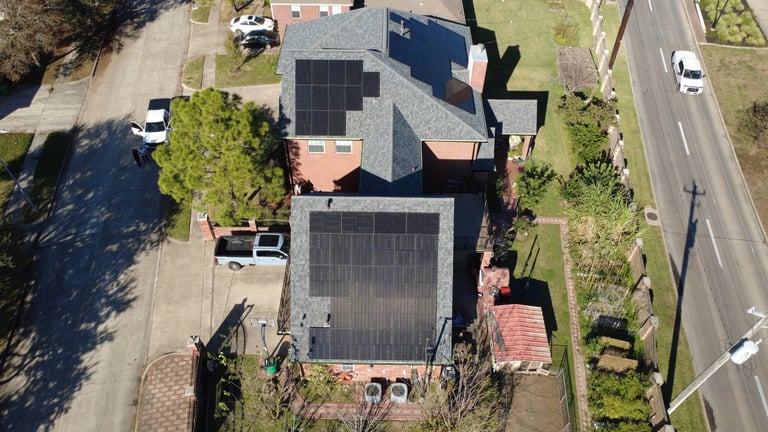 Aerial view of a residential house with solar panels installed on the roof and driveway, surrounded by trees and lawn