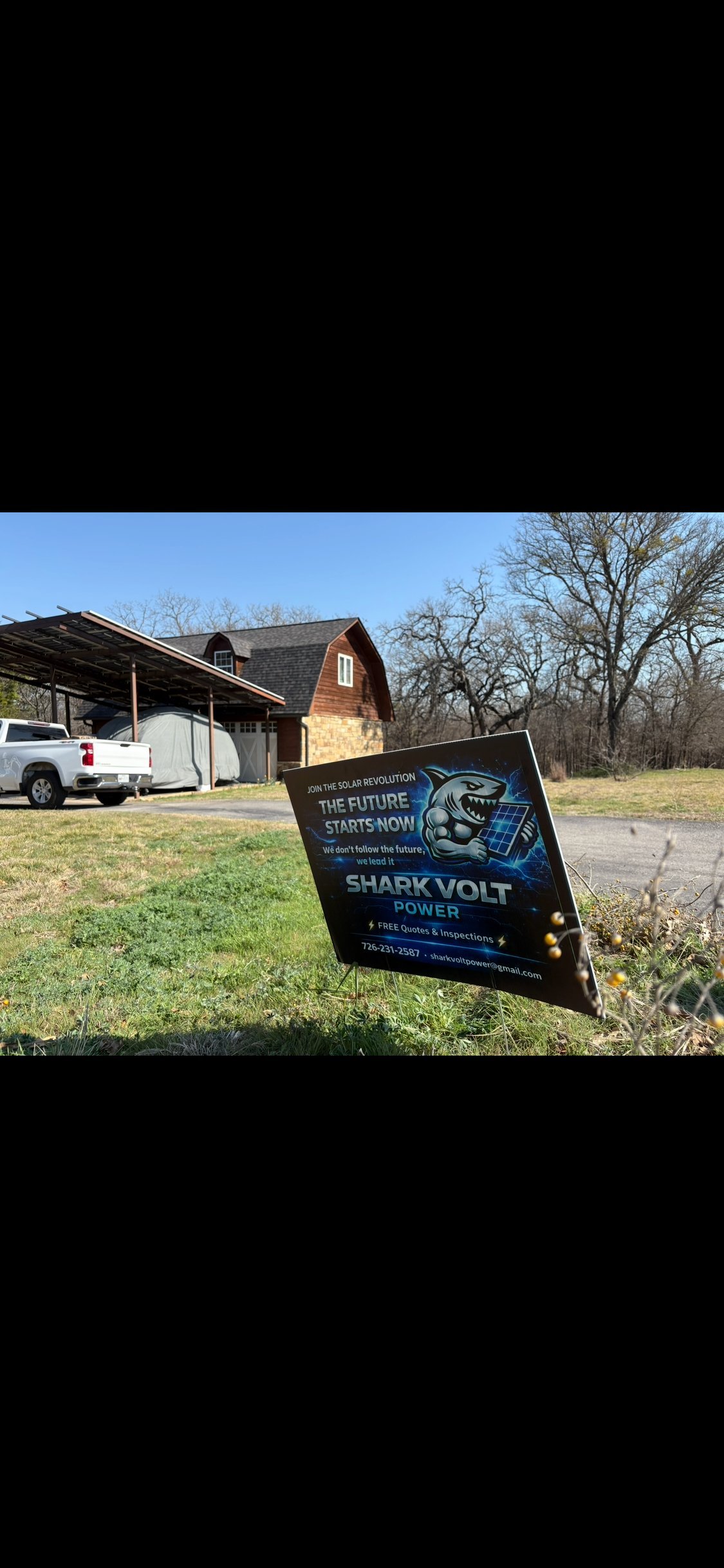 A Shark Volt Power yard sign on a residential property with a brick house, metal carport, and white truck visible in the background