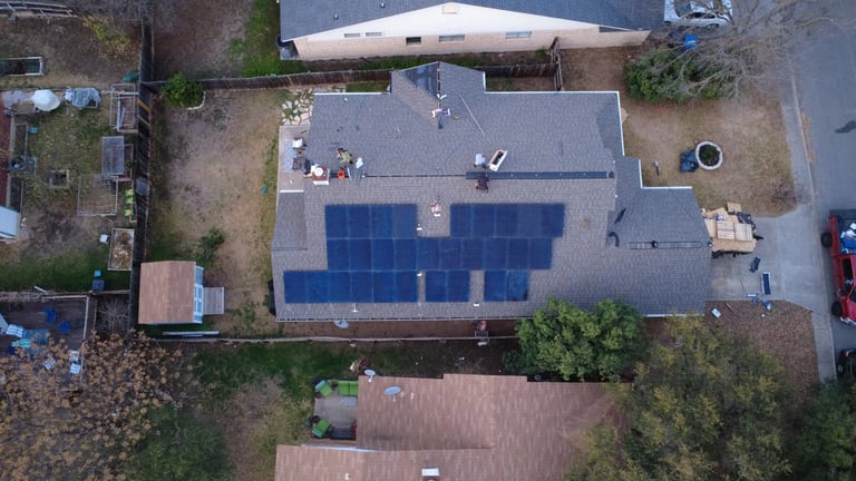 Aerial view of a residential roof with blue solar panels installed, surrounded by landscaping and neighboring properties