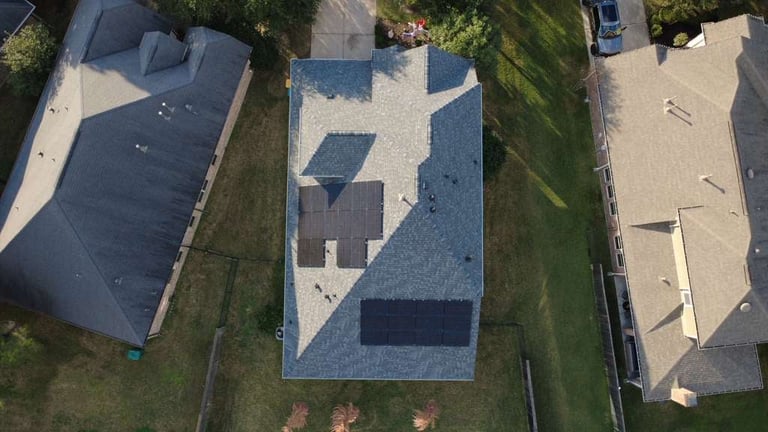 Aerial view of three residential houses with different roof types: dark asphalt shingle, blue composite tiles, and light beige roof, separated by green lawn strips