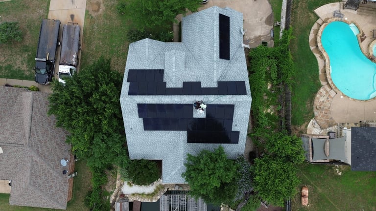 Aerial view of a modern white house with black roof panels surrounded by green lawn and neighboring properties with a blue pool visible